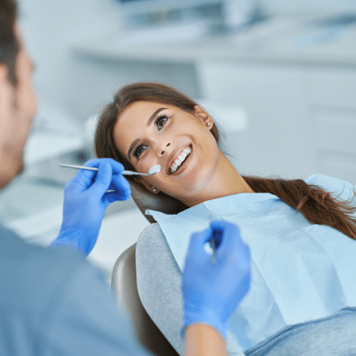 A dentist wearing blue gloves is attending to a patient lying back in a dental chair, with dental office equipment in the background.